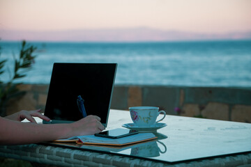 Woman remotely working from laptop and writing notes seaside on holiday. 