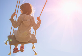 Back view of carefree happy young girl on swing in sunlight against blue sky. Copy space for text or design. - obrazy, fototapety, plakaty