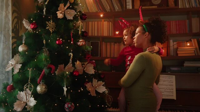 Mom holding daughter and standing in their living room next to the christmas tree and decorating it.