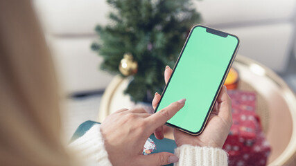 Over the shoulder of young blond woman looking and swiping smartphone with greenscreen and christmas decoration around