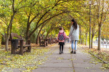 Mom and daughter walk in autumn park. Little girl after school with mother from back. Everyday life