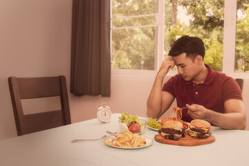 Concept of reducing junk food for health : handsome guy wants to have lunch, but the table is full of junk food such as french fries, hamburgers, and he pondered to control the food for good health. - obrazy, fototapety, plakaty