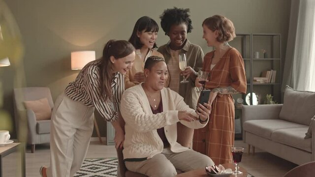 Medium long of short-haired Biracial woman sitting on chair in living room, holding smartphone, taking selfies with four young multiethnic female friends at daytime