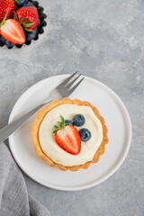 Delicious mini tarts with fresh berries and custard on a white plate on a gray concrete background. Copy space