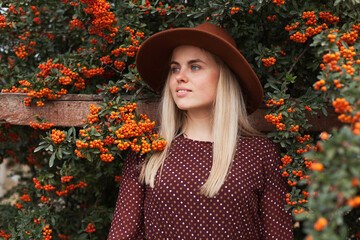 Beautiful woman wearing terracotta brown colous hat and rustic dress near rowan tree with ripe orange berries. Slow living and natural beauty concept.