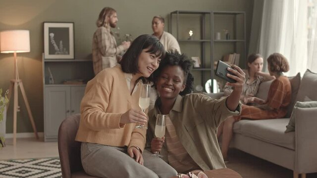 Medium long of young Asian and African American women holding glasses with cocktail, taking selfie with smartphone in living room at daytime, blurred people hanging out on background