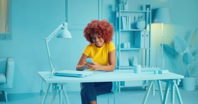 Slow motion of pretty young black woman with afro hairstyle holding smart phone and checking social media app while sitting in studio with blue interior. Curly female model using mobile phone at home