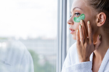 young woman in bathrobe applying green eye patch near window