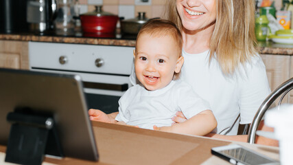 Happy mother and child using tablet together while sitting in kitchen at home. Laughing family, mom...