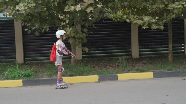 Little girl learns to roller skate. Kid is dressed in protection and a helmet, a schoolgirl rides along the road on roller skates