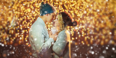 Having fun together at a christmas fairy with snowfall. Young cheerful couple is having a walk with hot drinks, enjoying, dressed warm, looking at each other and laugh, snowflakes all around