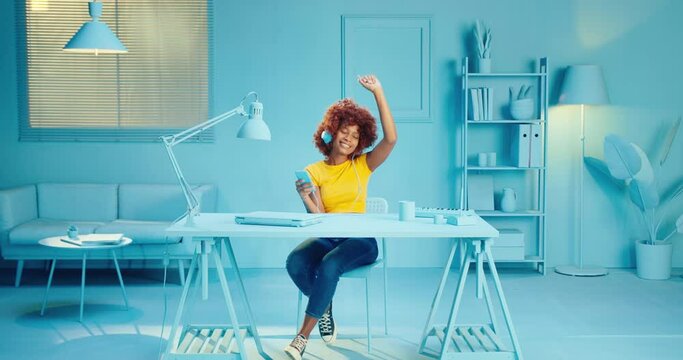 Slow motion of happy young black woman in casual yellow t shirt dancing and enjoying music on smart phone app with headphones at studio with blue interior. Carefree girl listening song on phone