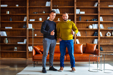 Fototapeta premium Two young smiling cheerful positive cool male friends raised their thumbs up together at home. People lifestyle concept