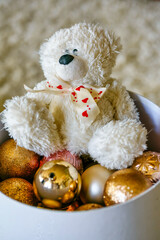 An adorable white teddy bear sits in a box with golden Christmas balls. Soft plush toy, gifts for the holidays. Decoration for a celebration or greeting cards. Romantic talisman, tenderness and love.