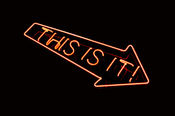 Neon sign in red with text saying THIS IS IT. Downward pointing arrow isolated on black