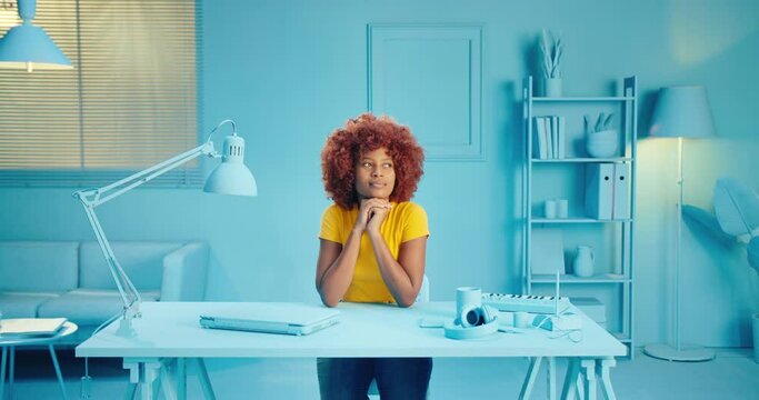 Portrait of pensive young African American woman with Afro hairstyle thinking looking up with hands on chin and smiling happy in studio. Curly girl with thinking pose and sitting at home