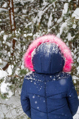 Rear view of little happy girl in pink warm fur hood walking outside on nature winter snowy forest background. Pretty child outdoor, cold weather. Caucasian kid. Winter nature. Vertical. Back view