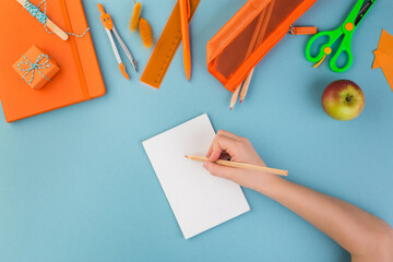 Child hand writes on white peace of paper school or office workspace with orange supplies on blue background. Flat lay.