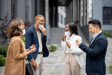 Multiracial business people taking off medical masks and talking on city street. Concept of health protection during Coronavirus pandemic. Joyful businessmen and businesswomen wearing formal wear