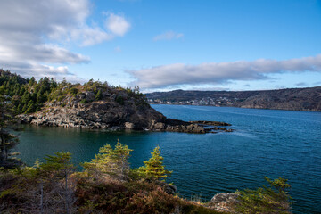 A rugged coastline in the Atlantic Ocean. The rugged cliffs of rocks have some vegetation and trees. It's a bright sunny day with a blue sky and few clouds. The ocean is deep blue with some waves.