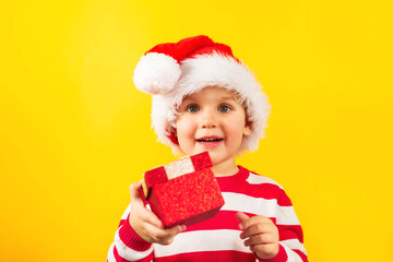 Funny child toddler in a Santa hat with red gift , yellow background. Holidays, Traditions, Christmas concept.