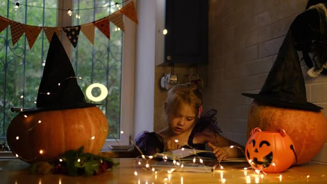 little girls in witch costume are reading fairy tale book with pictures. . Preparing for Halloween at home kitchen. Scary and fun holiday with family.