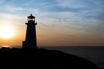 Lighthouse tower made of white concrete hexagonal, six-sided tapering concrete. The structure stands on a rock cliff. The sky has an orange color sunset. There's a green lantern in the cupola. 