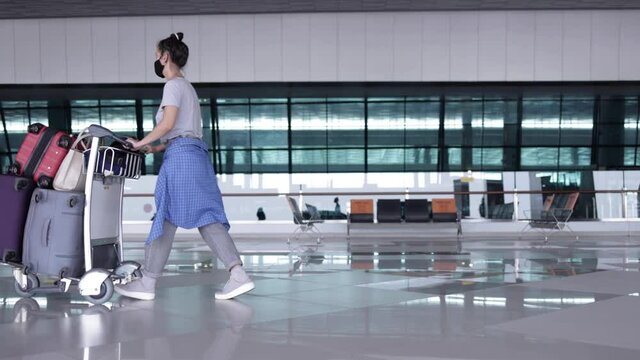 Young woman with face mask walking at airport terminal pushing luggage trolley with many bags before check in. Pandemic and traveling concept