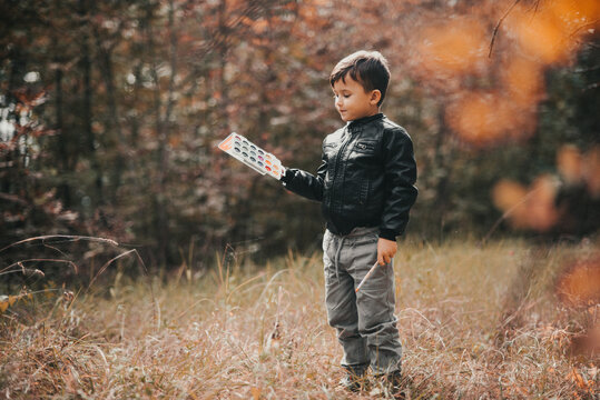A little boy of five years old is standing in the forest with paints in his hands, trying to paint the leaves of trees with paints