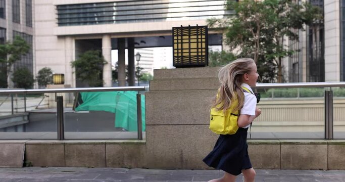 Happy little caucasian blonde girl seven years old in uniform with yellow backpack running back to school after summer vacations or Coronavirus covid-19 quarantine. 4k slow motion