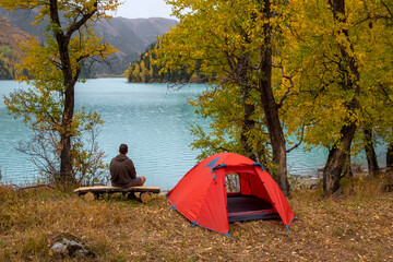Tourist is sitting near tent on the shore of beautiful lake with turquoise water and colorful forest around it. Tourism, travel, hiking in autumn season concept. Zhasylkol lake in Dzungarian Alatau.