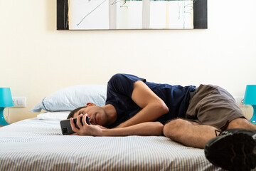 Bored young man lying on a bed in a bright room using a cell phone.