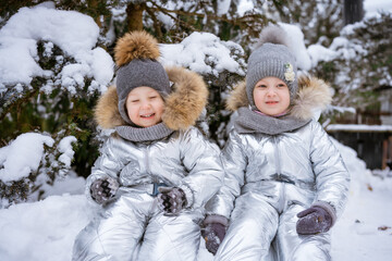 Fototapeta na wymiar Little brother and sister on winter walk in park in winter overalls sit in snow against background of snow-covered trees in knitted hats and a scarf. Happy childhood winter holidays and holidays
