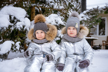 Fototapeta na wymiar Little brother and sister on winter walk in park in winter overalls sit in snow against background of snow-covered trees in knitted hats and a scarf. Happy childhood winter holidays and holidays
