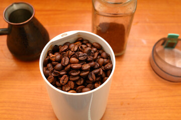 Electric coffee grinder with white plastic case, open transparent lid ready to grind fresh aromatic roasted coffee beans on background of brown table top with turk, glass jar closeup