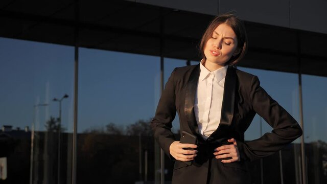 Business woman using smartphone touching stomach, feeling abdominal pain, indigestion or constipation because of unhealthy eating, symptoms of ulcer or gastritis because of nerves, illness outdoors.