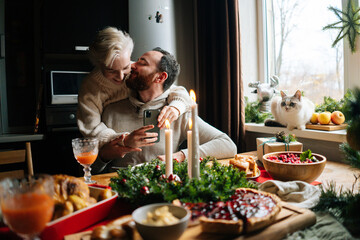 Portrait of happy young couple hugging sitting at festive Christmas table during holiday family party. Cheerful female taking photo or making video of beautiful xmas dinner table on mobile phone.