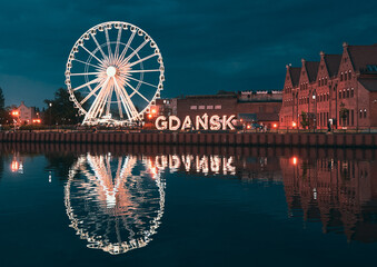 Reflection of the Ferris wheel in Gdansk, Poland. Old town center in the touristic city on the shore of the Baltic Sea in Poland. Beautiful night city lights - obrazy, fototapety, plakaty