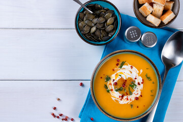 Butternut squash soup with cream, croutons, pumpkin seeds and blue napkin on one white wooden table. Copy space. Top view