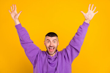 Photo of funky young brunet guy hands up wear purple hoodie isolated on yellow color background