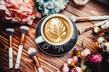 Cup of coffee on wooden brown background with flowers and makeup brushes