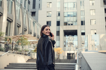 Cheerful young brunette woman talking on mobile phone smiling toothy smile looking over shoulder at camera climbing stairs to modern office corporate high-rise building.