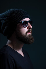 Obraz premium authentic portrait of a bearded millennial in sunglasses and a black knitted hat against a dark background