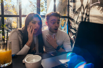 Cute couple are looking surprised at their laptop while sitting in a urban cafe near a window