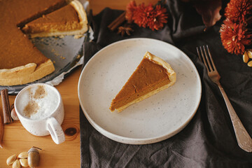 Happy Thanksgiving. Pumpkin pie slice on modern plate and hot coffee on rustic table with linen napkin, autumn flowers and leaves, anise and cinnamon. Homemade pumpkin tart recipe.