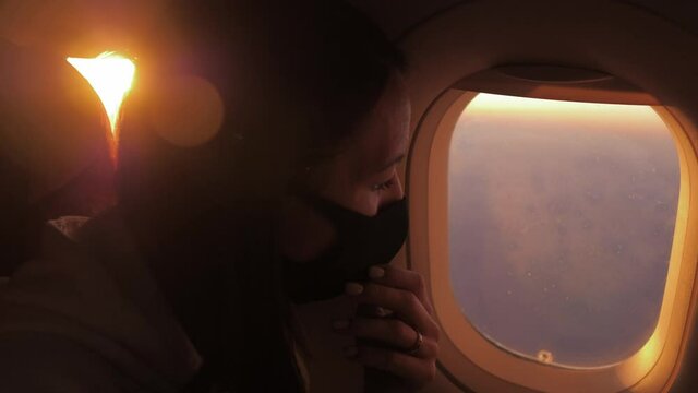 Young woman sits on a plane in a black medical respiratory mask and looks out the window. Health, travel concepts. Safety and new rules of conduct for air transport passengers.