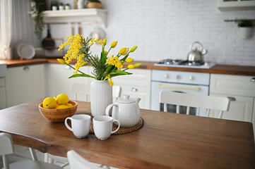 Fototapeta na wymiar a bouquet of yellow tulips and mimosa in a vase on a wooden table against the background of a white modern kitchen, white dishes and lemons.
