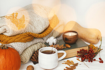 Pumpkin, cup of tea, cozy sweaters, autumn leaves, burning candle and vintage book in golden lights bokeh on white wooden background in room. Hello autumn, cozy slow living. Happy Thanksgiving