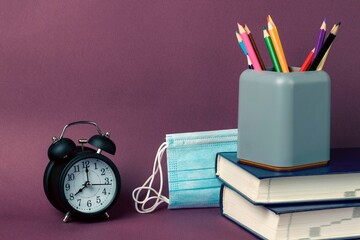Black alarm clock, a stack of books, multicolored pencils in a glass and medical masks on a purple...
