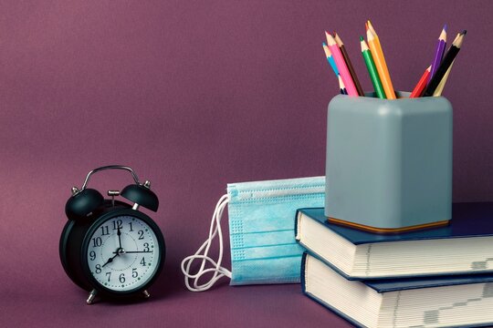 Black alarm clock, a stack of books, multicolored pencils in a glass and medical masks on a purple background. Back to school, coronavirus restrictions